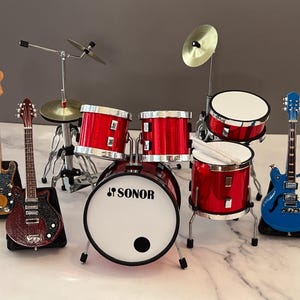 May include: Miniature red drum kit with a SONOR logo, cymbals, and drumsticks. Includes two guitars, one yellow and one burgundy, and a blue guitar. A fun musical instrument set.
