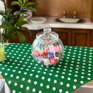 May include: A glass jar filled with colourful lollipops sits on a green tablecloth with white polka dots. A clear glass vase with a white flower and yellow beads is on the table.
