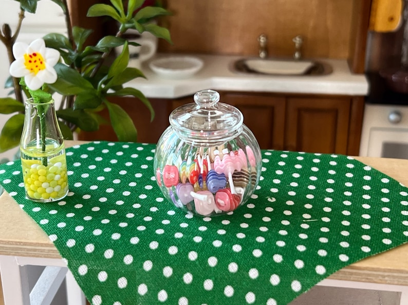 May include: A glass jar filled with colourful lollipops sits on a green tablecloth with white polka dots. A clear glass vase with a white flower and yellow beads is on the table.