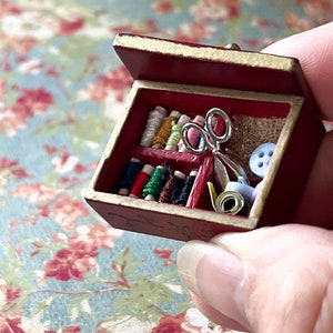 Puede incluir: Una peque&ntilde;a caja de madera roja con tapa abatible, llena de carretes de hilo de colores, un par de tijeras, un rollo de cinta y un bot&oacute;n.