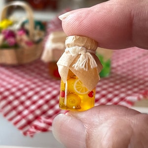 Może przedstawiać: Miniaturowy szklany słoik wypełniony żółtymi plastrami cytrusów i czerwonymi jagodami, przykryty brązową papierową pokrywką i białym sznurkiem. Słoik jest trzymany przez rękę.