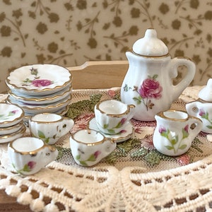 May include: A miniature tea set with a white teapot, creamer, and sugar bowl with pink rose floral designs. The set includes four teacups and saucers with matching floral designs. The tea set is displayed on a white crocheted doily.