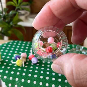 May include: A clear glass jar filled with colourful miniature lollipops. The jar is being held by a hand, and there are more lollipops scattered on a green and white polka dot surface.