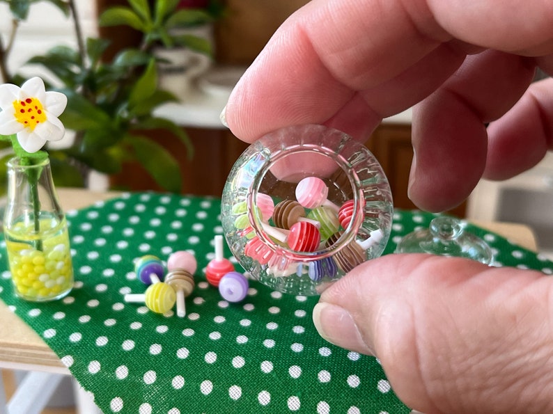 May include: A clear glass jar filled with colourful miniature lollipops. The jar is being held by a hand, and there are more lollipops scattered on a green and white polka dot surface.