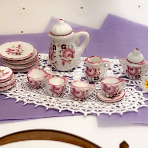 May include: A miniature tea set with a white teapot, creamer, sugar bowl, and four teacups and saucers. The set is decorated with pink roses and green leaves.