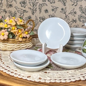 May include: A miniature white ceramic dish set with a basket of yellow and pink flowers. The set includes small plates, a pitcher with green leaf designs, and a decorative lace doily.