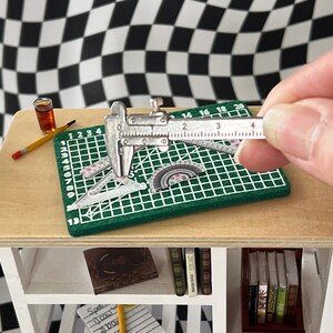 May include: A close-up of a silver caliper measuring a green cutting mat with a ruler and protractor. The mat has a grid pattern and numbers 1-20. The caliper has a black handle and a silver measuring scale.