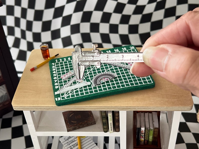 May include: A close-up of a silver caliper measuring a green cutting mat with a ruler and protractor. The mat has a grid pattern and numbers 1-20. The caliper has a black handle and a silver measuring scale.