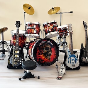 May include: A miniature drum kit and guitars with a Metallica theme. The drum kit features a skull graphic and flame design. Guitars include black, silver, and blue colours. The set includes a drum stool and foot pedal.