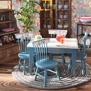 Puede incluir: Un juego de comedor en miniatura con una mesa azul y blanca y cuatro sillas azules. La mesa tiene un mantel blanco con estampado floral y una jarra de limonada rosa. Las sillas tienen respaldos con estampado floral. El juego está sobre una alfombra marrón con un patrón blanco y gris.