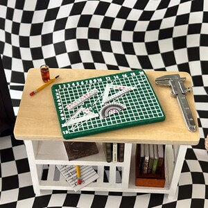 May include: A white wooden shelf with a green cutting mat, rulers, a protractor, a pencil, a glass of liquid, and a silver caliper. The shelf has two shelves and is sitting on a black and white checkered surface.