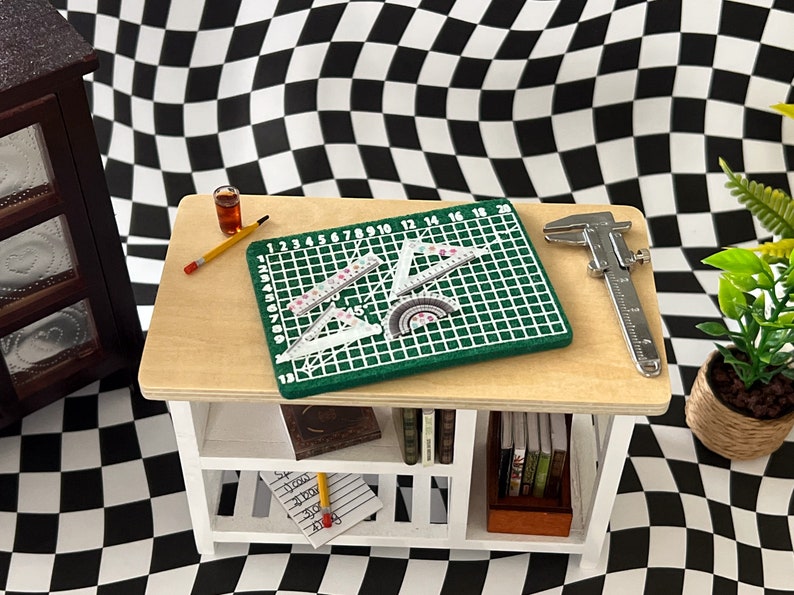 May include: A white wooden shelf with a green cutting mat, rulers, a protractor, a pencil, a glass of liquid, and a silver caliper. The shelf has two shelves and is sitting on a black and white checkered surface.