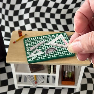 May include: A green cutting mat with a grid pattern and numbers 1-20. There are three clear plastic rulers on the mat, including a protractor. A hand is holding a clear plastic triangle ruler with a floral pattern.