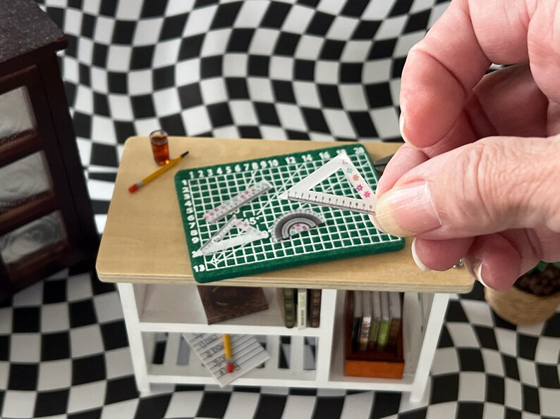 May include: A green cutting mat with a grid pattern and numbers 1-20. There are three clear plastic rulers on the mat, including a protractor. A hand is holding a clear plastic triangle ruler with a floral pattern.