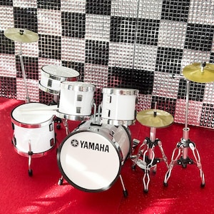 Op de afbeelding: Een witte Yamaha miniatuur drumstel met zilveren hardware en bekkens. De drumstel staat op een rode ondergrond, met een zwart-zilveren geblokte achtergrond. Het Yamaha-logo is zichtbaar op de grote trom.