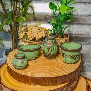May include: A collection of small, distressed green ceramic pots and vases with brown accents, arranged on a wooden slice. The set includes a small vase, a round pot, and two other decorative containers. The background features plants and flowers.