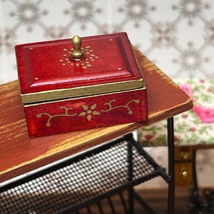 Puede incluir: Una peque&ntilde;a caja de madera roja con un pomo dorado en la tapa. La caja tiene dise&ntilde;os florales dorados pintados en la tapa y los lados. La caja est&aacute; sentada en una mesa de madera.