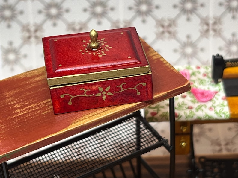 Puede incluir: Una peque&ntilde;a caja de madera roja con un pomo dorado en la tapa. La caja tiene dise&ntilde;os florales dorados pintados en la tapa y los lados. La caja est&aacute; sentada en una mesa de madera.