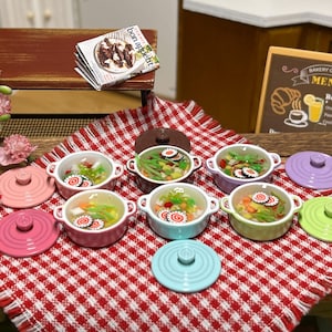 Könnte beinhalten: Ein Set aus neun Miniatur-Keramiktöpfen mit Deckeln, die jeweils mit bunten, simulierten Lebensmitteln gefüllt sind. Die Töpfe sind auf einer rot-weiß karierten Tischdecke angeordnet.