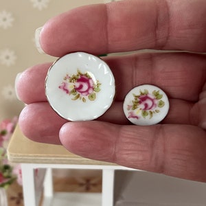 Może przedstawiać: Dwa miniaturowe talerze porcelanowe z wzorem r&oacute;żowej r&oacute;ży. Talerze są białe z pozłacanymi krawędziami i przedstawiają r&oacute;żową r&oacute;żę z zielonymi liśćmi.