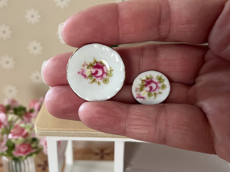 Może przedstawiać: Dwa miniaturowe talerze porcelanowe z wzorem r&oacute;żowej r&oacute;ży. Talerze są białe z pozłacanymi krawędziami i przedstawiają r&oacute;żową r&oacute;żę z zielonymi liśćmi.