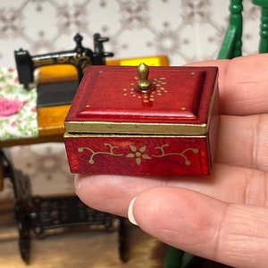 Puede incluir: Una peque&ntilde;a caja de madera roja con tapa dorada y dise&ntilde;os florales pintados en oro. La caja tiene un peque&ntilde;o pomo dorado en la parte superior.