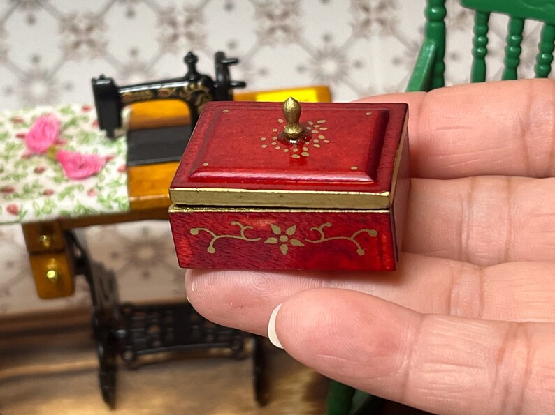 Puede incluir: Una peque&ntilde;a caja de madera roja con tapa dorada y dise&ntilde;os florales pintados en oro. La caja tiene un peque&ntilde;o pomo dorado en la parte superior.