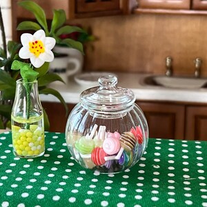 May include: A clear glass jar with a lid filled with colourful sweets. The jar is sitting on a green tablecloth with white polka dots. A vase with a white flower and yellow and green beads is in the background.