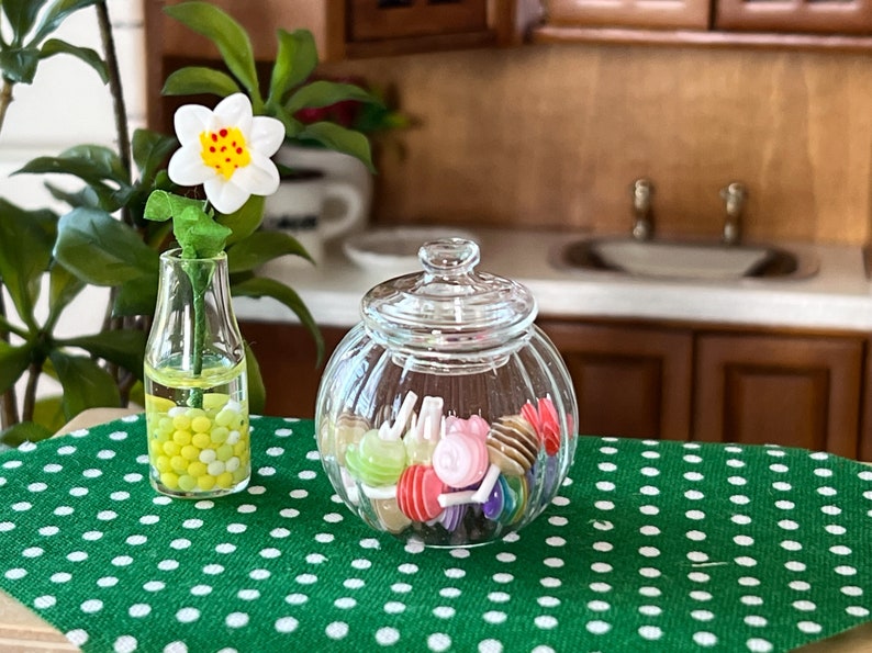 May include: A clear glass jar with a lid filled with colourful sweets. The jar is sitting on a green tablecloth with white polka dots. A vase with a white flower and yellow and green beads is in the background.