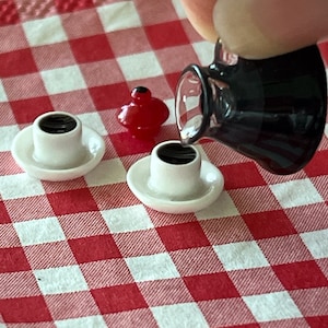 May include: A miniature coffee pot pouring coffee into a miniature white coffee cup on a red and white checkered tablecloth. There is a red miniature bird figurine on the tablecloth.