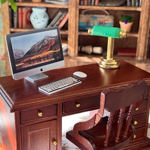 Puede incluir: Un escritorio de madera en miniatura con una computadora, un teclado, un ratón y una lámpara de escritorio verde. El escritorio tiene tres cajones y una puerta de armario. Una silla de madera está colocada a la derecha del escritorio.