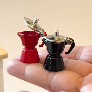 May include: Two miniature espresso makers, one red and one black, with silver accents. The red espresso maker has its lid open.