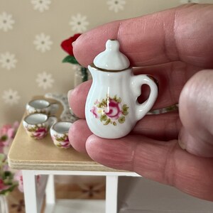 Może przedstawiać: Miniaturowa biała porcelanowa czajniczka z kwiatowym wzorem przedstawiającym r&oacute;żowe r&oacute;że i zielone liście. Czajniczek ma pokrywkę i uchwyt.