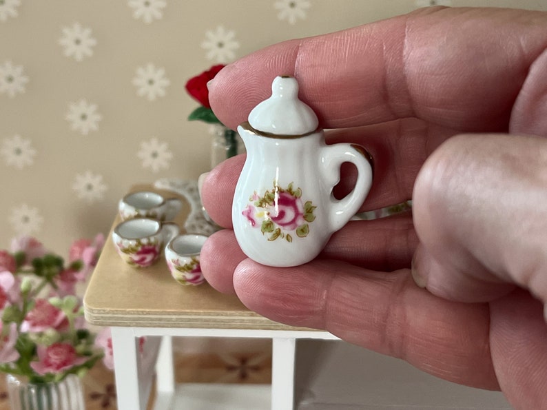 Może przedstawiać: Miniaturowa biała porcelanowa czajniczka z kwiatowym wzorem przedstawiającym r&oacute;żowe r&oacute;że i zielone liście. Czajniczek ma pokrywkę i uchwyt.