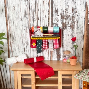 Puede incluir: Un estante de madera en miniatura con ribete rojo contiene carretes de cinta y una regla pequeña. El estante está montado en un panel de madera blanca. Un camino de mesa de tela roja está sobre una mesa de madera con un rollo de tela blanca y un frasco de vidrio de cuentas de colores.