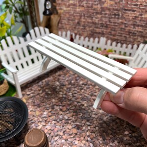 Peut inclure: Banc miniature en bois blanc avec quatre lattes. Le banc est tenu dans une main, avec une cl&ocirc;ture en bois blanc et un jardin miniature en arri&egrave;re-plan.