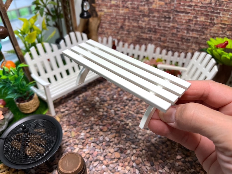 Peut inclure: Banc miniature en bois blanc avec quatre lattes. Le banc est tenu dans une main, avec une cl&ocirc;ture en bois blanc et un jardin miniature en arri&egrave;re-plan.