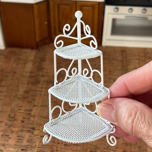 Könnte beinhalten: Ein Miniatur-Eckregal aus weißem Metall mit drei Ebenen. Die Regale sind dreieckig mit einem gewebten Muster. Der Rahmen hat dekorative Schnörkel und einen Kugelaufsatz. Eine Hand hält das Regal.