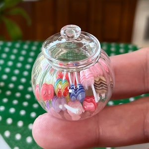 May include: A clear glass jar filled with colourful candy lollipops. The lollipops are in various shapes and sizes, including hearts, stars, and swirls. The jar has a lid and is sitting on a green and white polka dot surface.