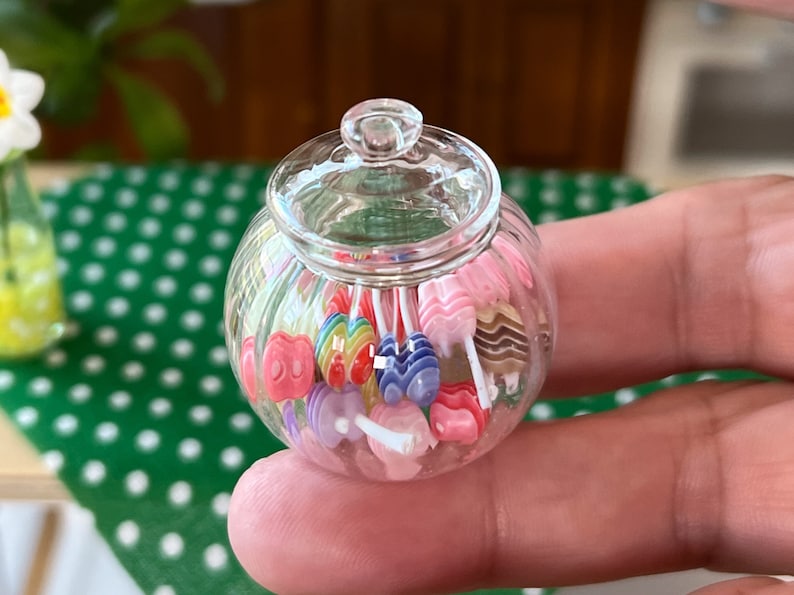 May include: A clear glass jar filled with colourful candy lollipops. The lollipops are in various shapes and sizes, including hearts, stars, and swirls. The jar has a lid and is sitting on a green and white polka dot surface.