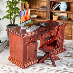Puede incluir: Miniatura de escritorio y silla de madera en un entorno de oficina en casa. El escritorio es de color marrón intenso con herrajes dorados. Un monitor de ordenador plateado, un teclado y un ratón se encuentran sobre el escritorio. Una estantería de madera está en el fondo.