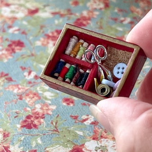 Puede incluir: Una caja de costura de madera en miniatura con un interior rojo. La caja est&aacute; llena de carretes de hilo de colores, un par de tijeras, un peque&ntilde;o trozo de tela y botones.