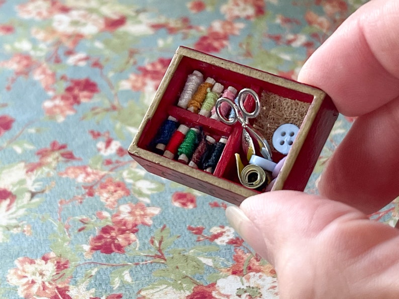 Puede incluir: Una caja de costura de madera en miniatura con un interior rojo. La caja est&aacute; llena de carretes de hilo de colores, un par de tijeras, un peque&ntilde;o trozo de tela y botones.