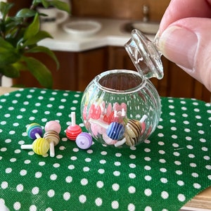 May include: A clear glass candy jar with a lid, filled with colourful candy lollipops. The jar is sitting on a green tablecloth with white polka dots. There are also some lollipops scattered around the jar.