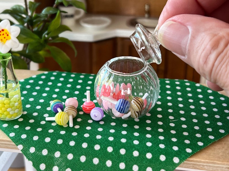 May include: A clear glass candy jar with a lid, filled with colourful candy lollipops. The jar is sitting on a green tablecloth with white polka dots. There are also some lollipops scattered around the jar.