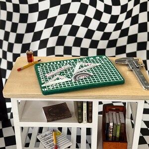May include: A white shelf unit with a green cutting mat, rulers, a pencil, a small glass bottle, and a silver caliper on the top shelf. The bottom shelves hold books and a notebook with the word "Spelling" written on it.