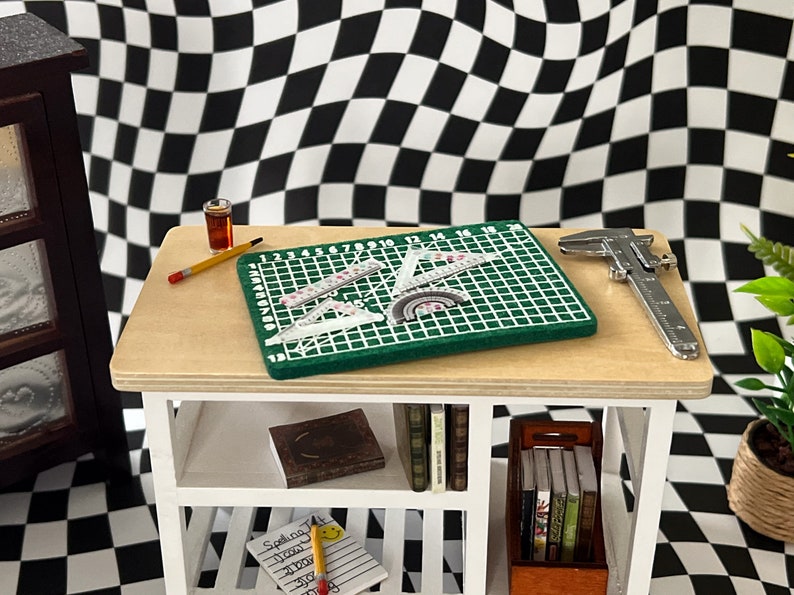 May include: A white shelf unit with a green cutting mat, rulers, a pencil, a small glass bottle, and a silver caliper on the top shelf. The bottom shelves hold books and a notebook with the word "Spelling" written on it.
