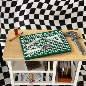 May include: A white wooden shelf unit with three shelves. The top shelf has a green cutting mat with rulers and protractors, a silver caliper, a yellow pencil, and a glass of brown liquid. The middle shelf has a book and the bottom shelf has a notepad and pencil.
