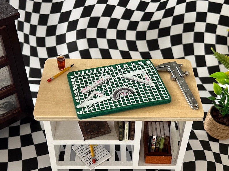May include: A white wooden shelf unit with three shelves. The top shelf has a green cutting mat with rulers and protractors, a silver caliper, a yellow pencil, and a glass of brown liquid. The middle shelf has a book and the bottom shelf has a notepad and pencil.