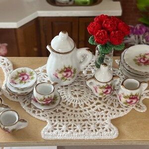 Może przedstawiać: Miniaturowy porcelanowy zestaw do herbaty z białym czajnikiem, dzbankiem na śmietanę, cukiernicą i czterema filiżankami i spodkami. Zestaw zdobiony jest kwiatowym wzorem z r&oacute;żowymi r&oacute;żami i zielonymi liśćmi. Zestaw do herbaty jest prezentowany na białym koronkowej serwetce z bukietem czerwonych r&oacute;ż w szklanym wazonie.